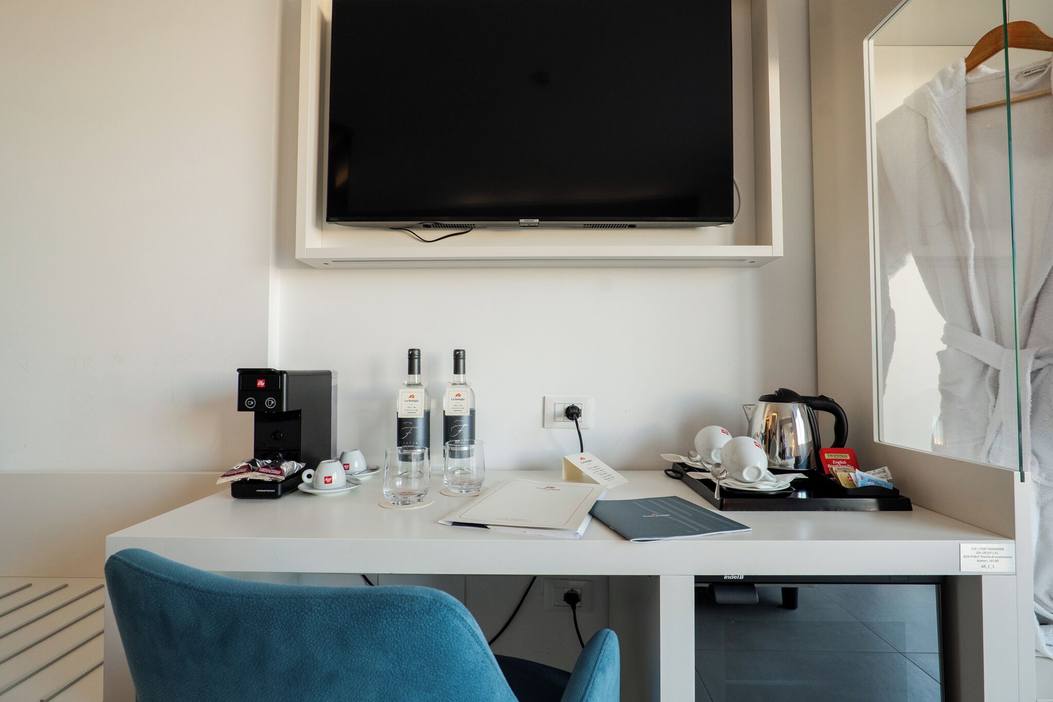 A hotel room with a white desk, a flat-screen TV, a coffee maker, two bottles of water, a notebook, and a laptop.