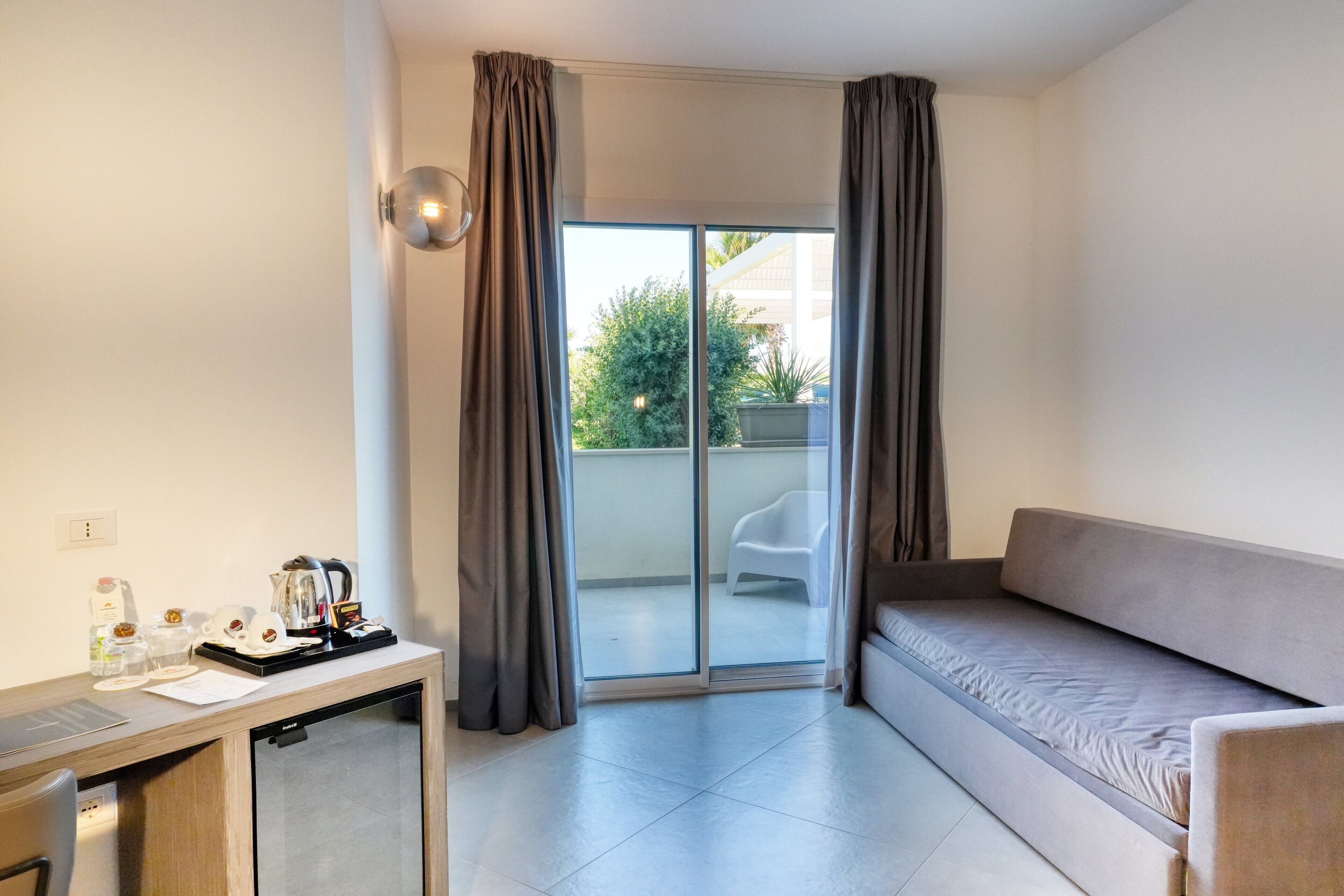 Een moderne kamer met een bank, een klein bureau met koffiezetapparaat, en een glazen schuifdeur naar een buitenruimte.