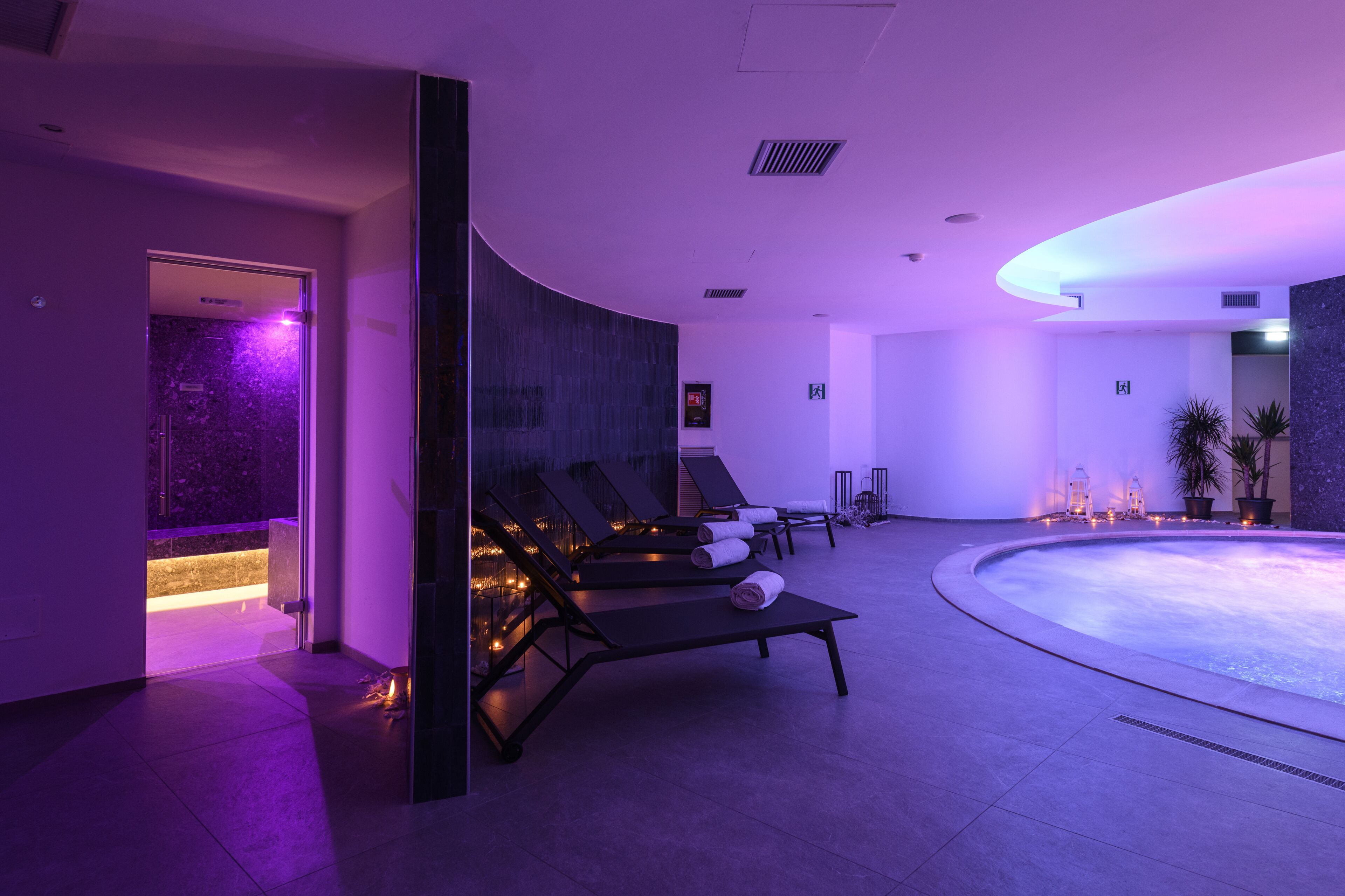Area piscina interna moderna con poltrona relax e piscina riscaldata, caratterizzata da un soffitto curvo e illuminazione d'ambiente.