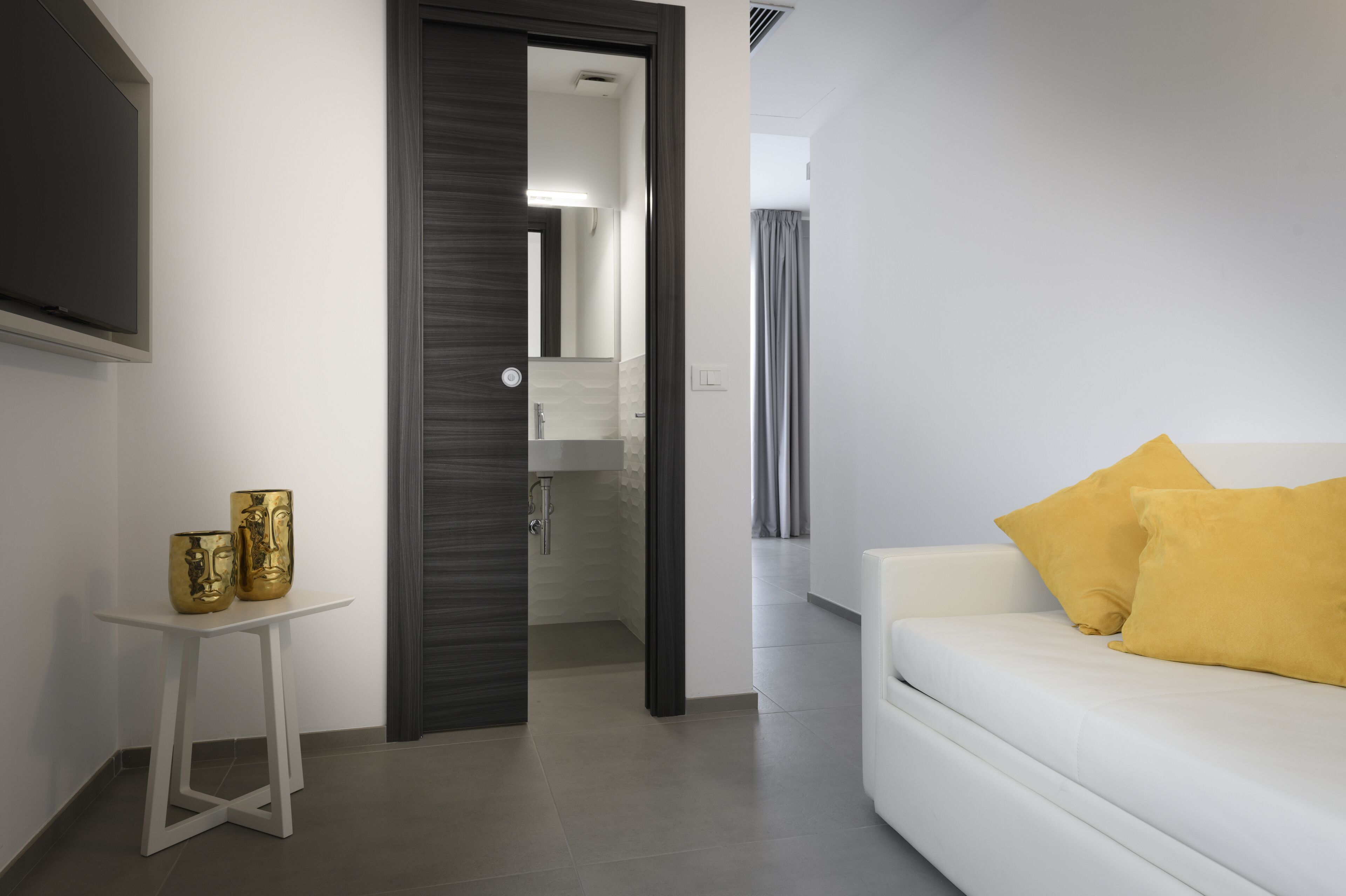A modern living room with a white sofa and yellow cushions, a small white table with decorative items, and a doorway leading to a bathroom.