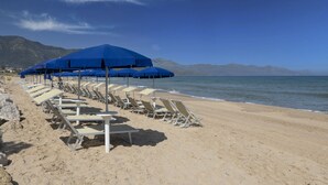 A beach with rows of sun loungers and blue umbrellas, a clear sky, and mountains in the background. - Resort La battigia Beach And Spa (Alcamo)
