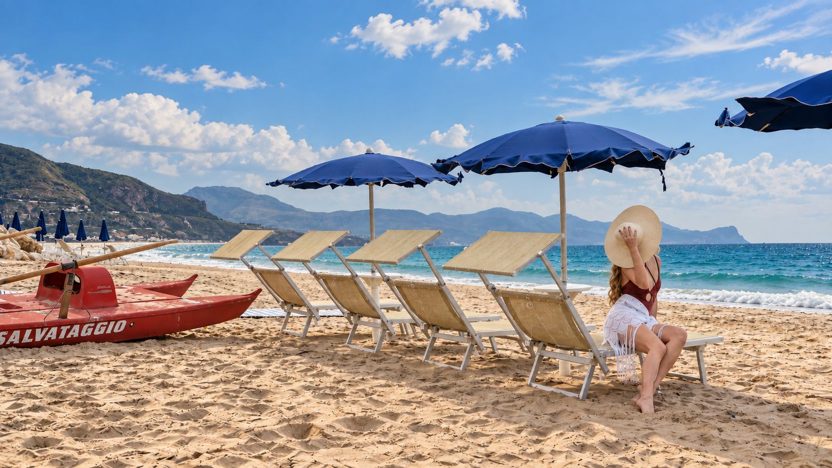 A beach with rows of sun loungers and umbrellas, a lifeguard boat, and a clear view of the sea and mountains.