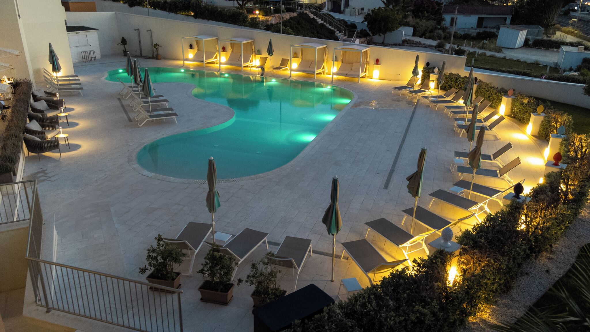 Zona de piscina con sillas de descanso y sombrillas, rodeada por un muro bajo y plantas en macetas.