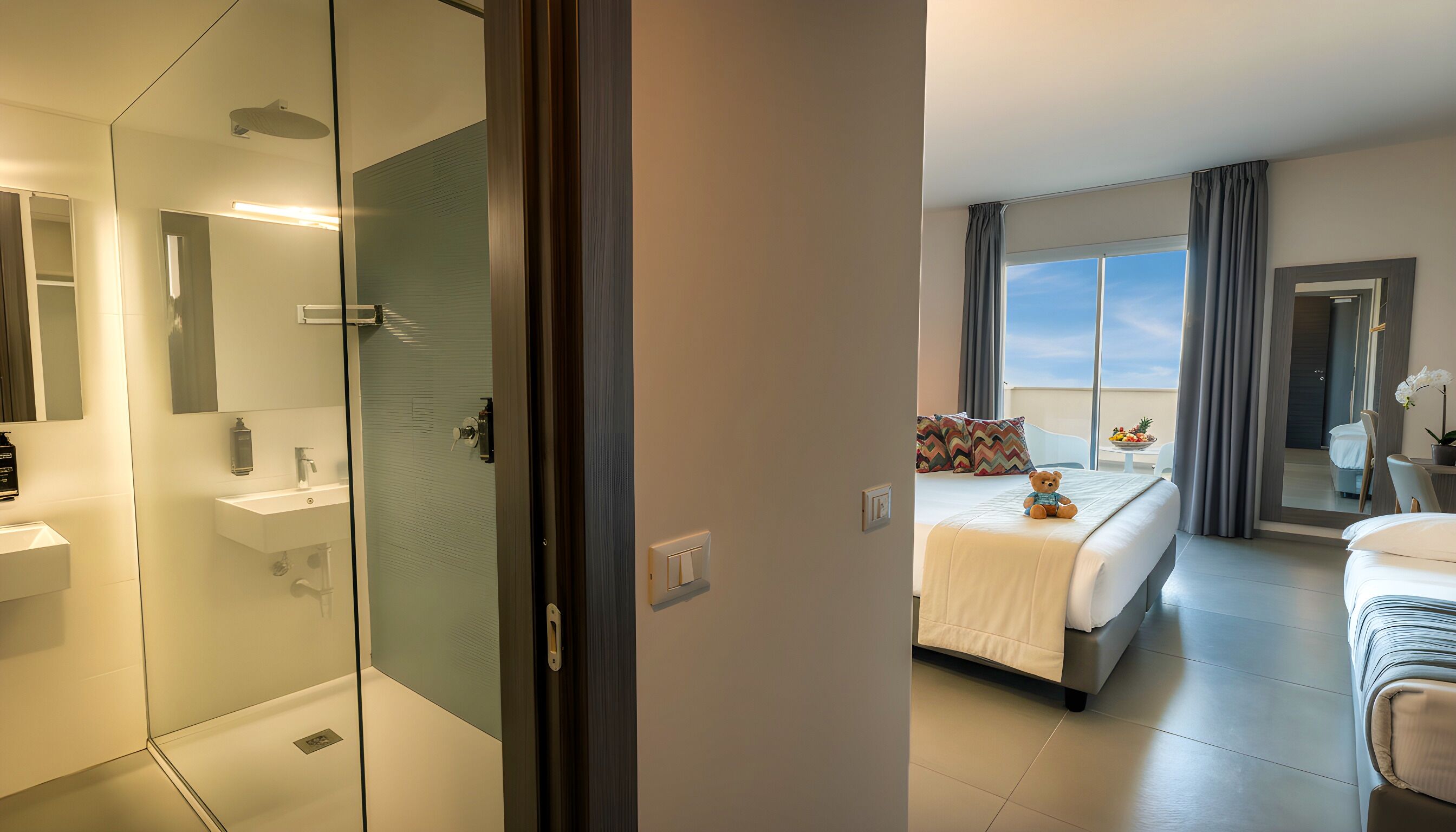 A modern hotel room with a glass shower enclosure, a separate bathroom, a large bed with a view of the sky, and a balcony with a fruit bowl.
