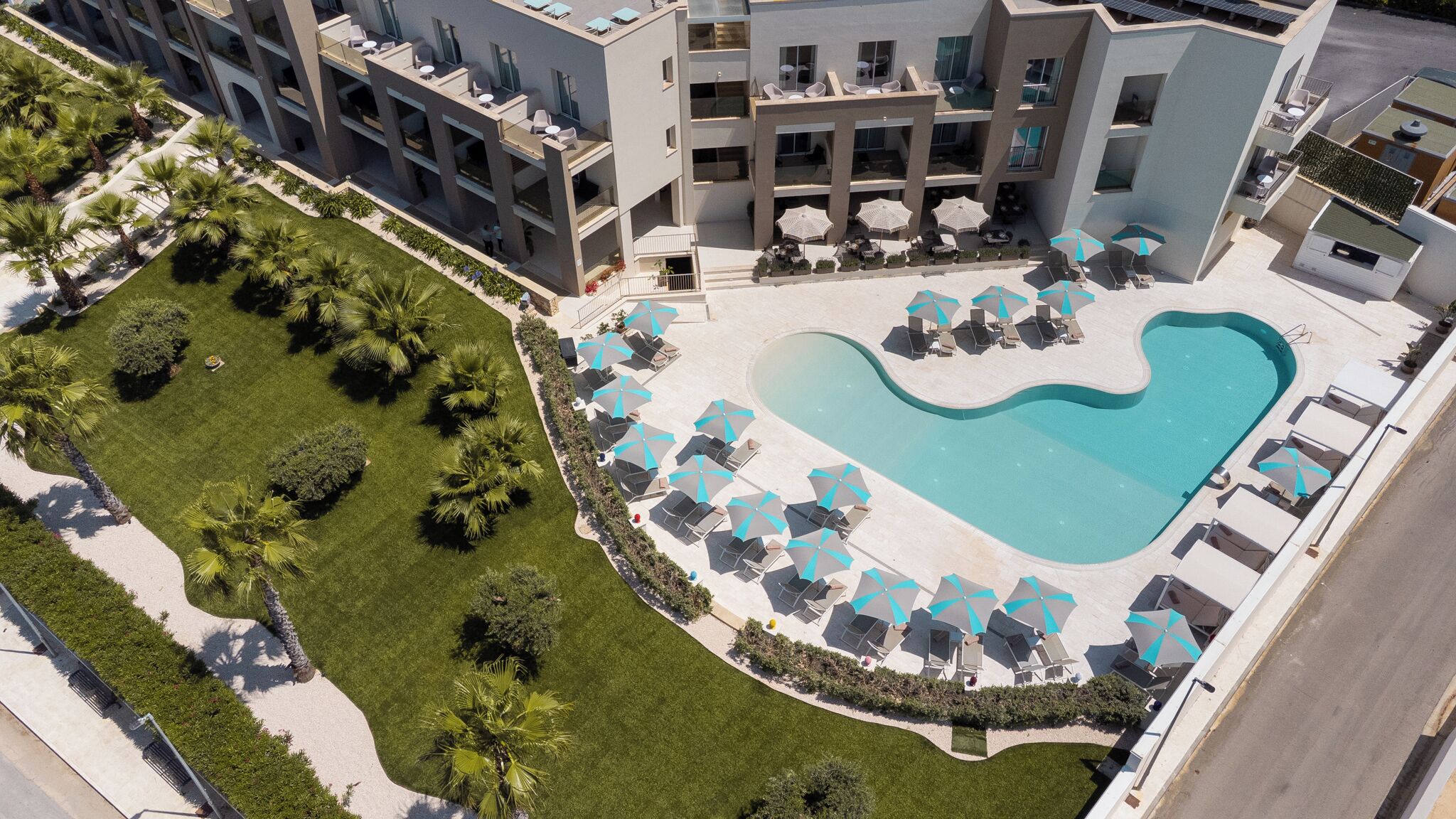 An aerial view of a modern hotel with a swimming pool, sun loungers, and a well-maintained garden.