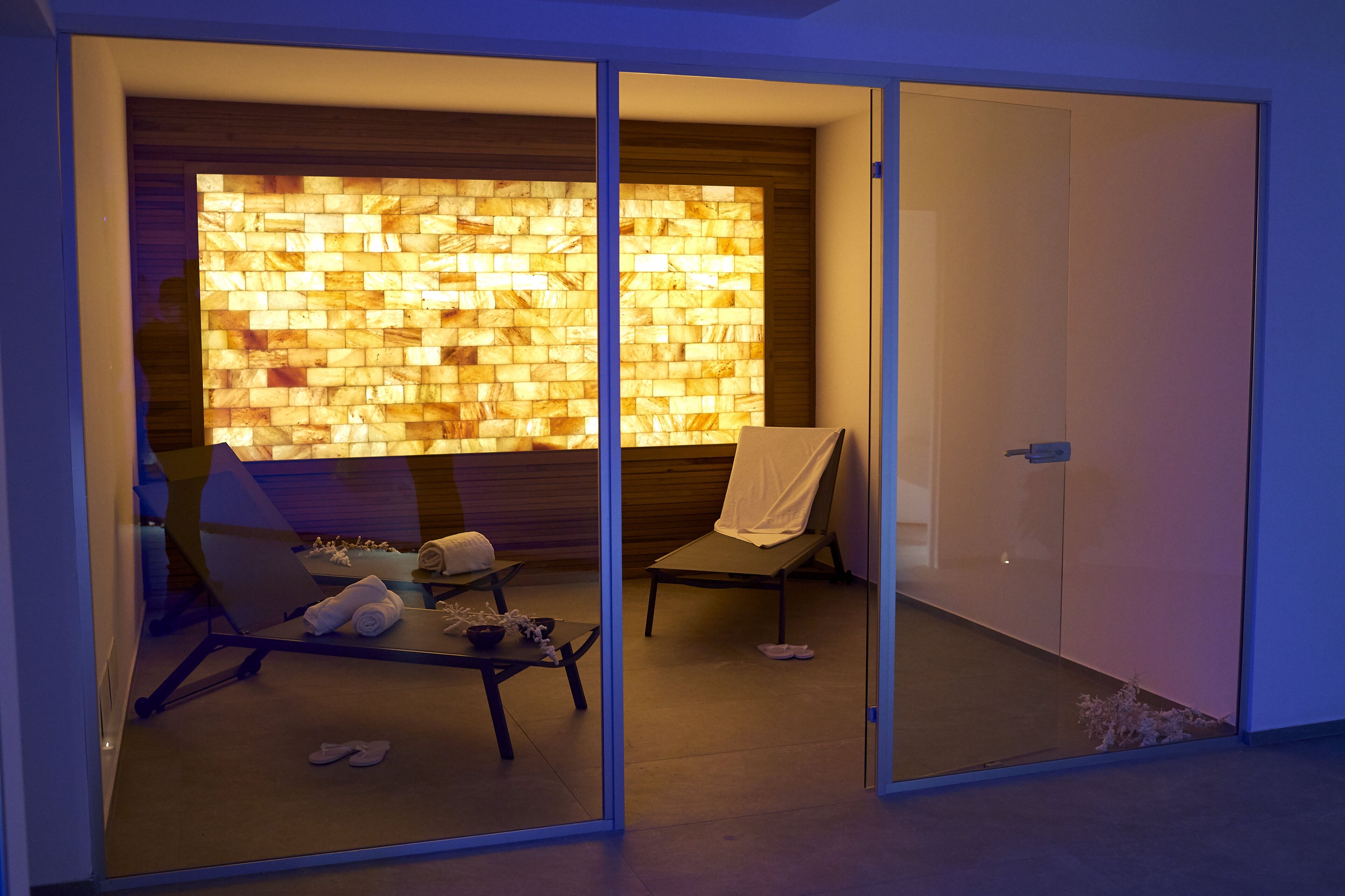 A modern room with a glass partition, a wooden wall with a light pattern, a lounge chair, a table with towels, and a door.