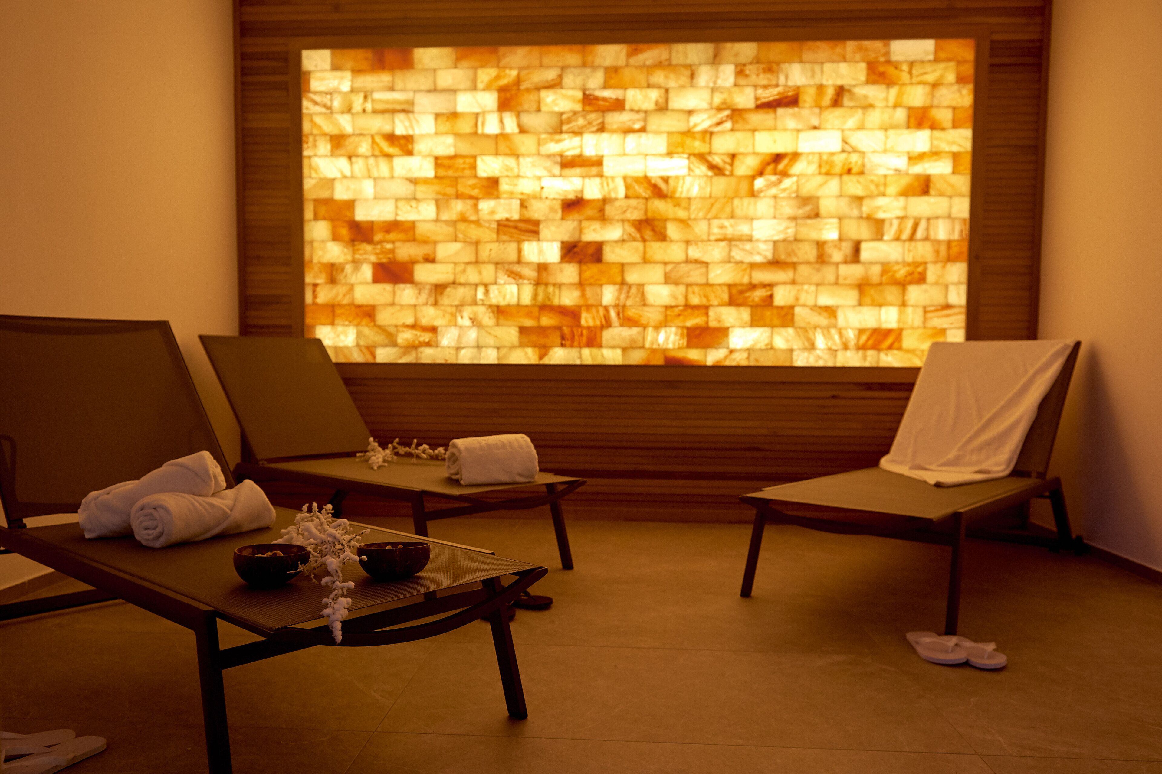 A spa room with three reclining chairs, each with a towel and a bowl. A large mosaic wall is visible in the background.