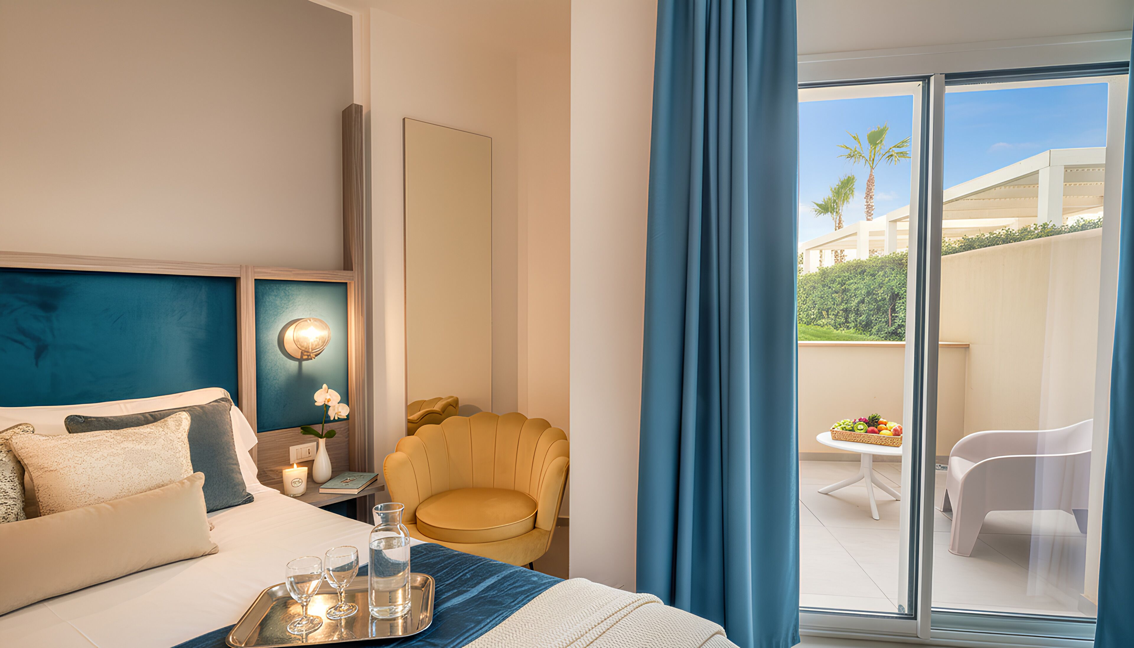 Een hotelkamer met een bed, een stoel, een tafel met fruit en een balkon met uitzicht op palmbomen.
