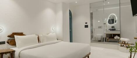 Un dormitorio moderno con una cama grande, mesitas de noche, un espejo y un baño visible a través de una puerta de cristal.