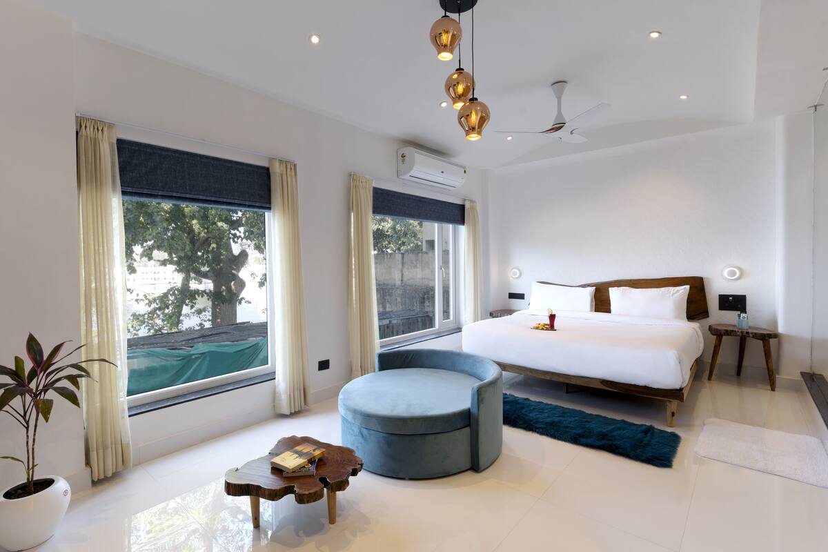 A modern hotel room with a large bed, a round ottoman, a small wooden table, and a potted plant.