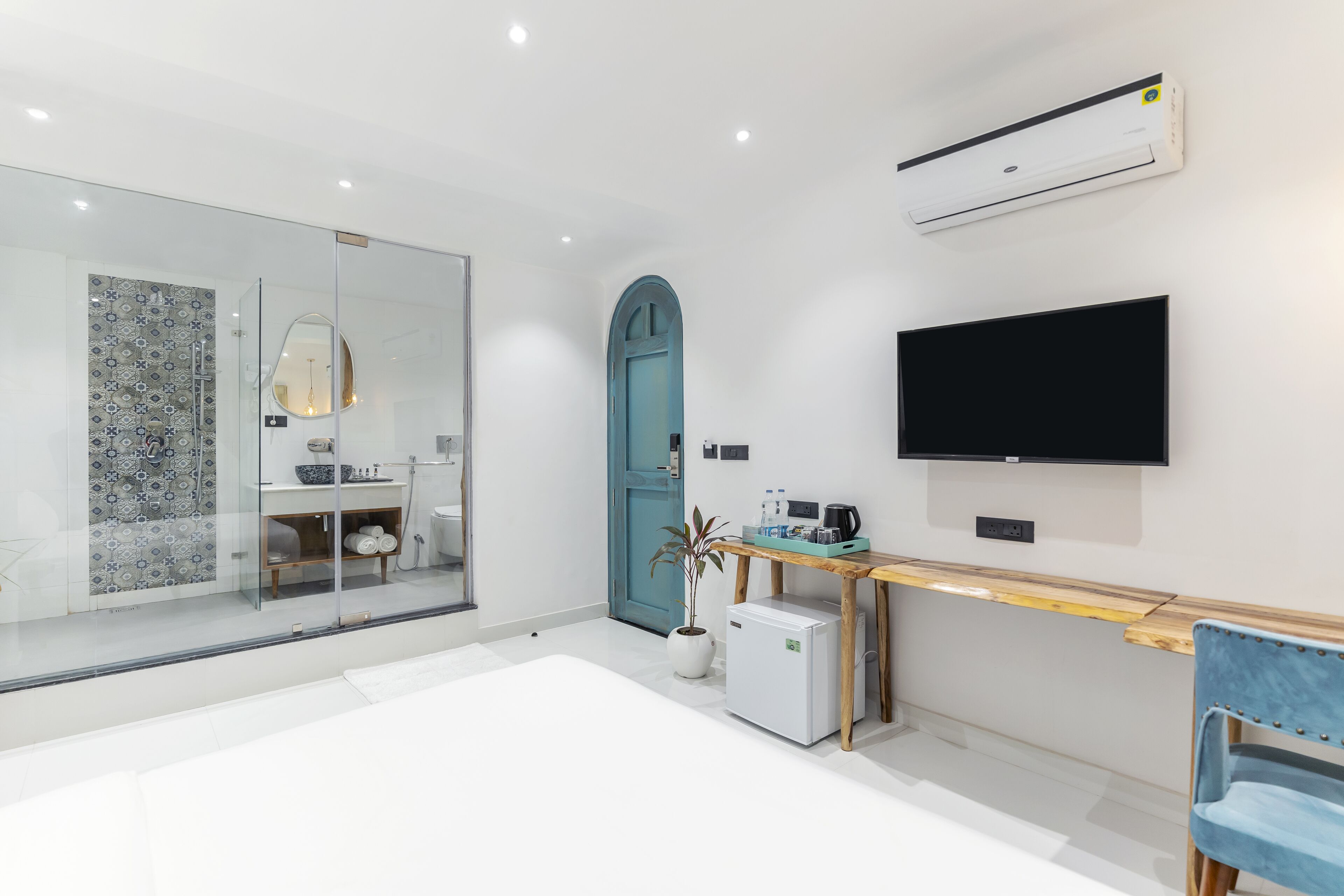 A modern hotel room with a glass shower, a teal door, a flat-screen TV, a small fridge, and a wooden table with a plant.