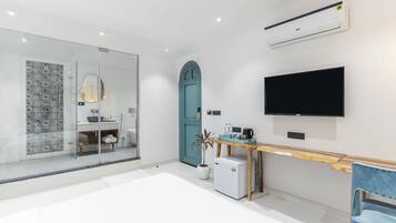 A modern hotel room with a glass shower, a teal door, a flat-screen TV, a small fridge, and a wooden table with a plant.