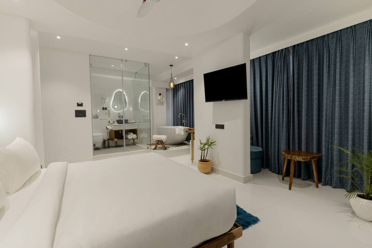 A modern bedroom with a large bed, a TV, a bathroom with a bathtub, and a potted plant.