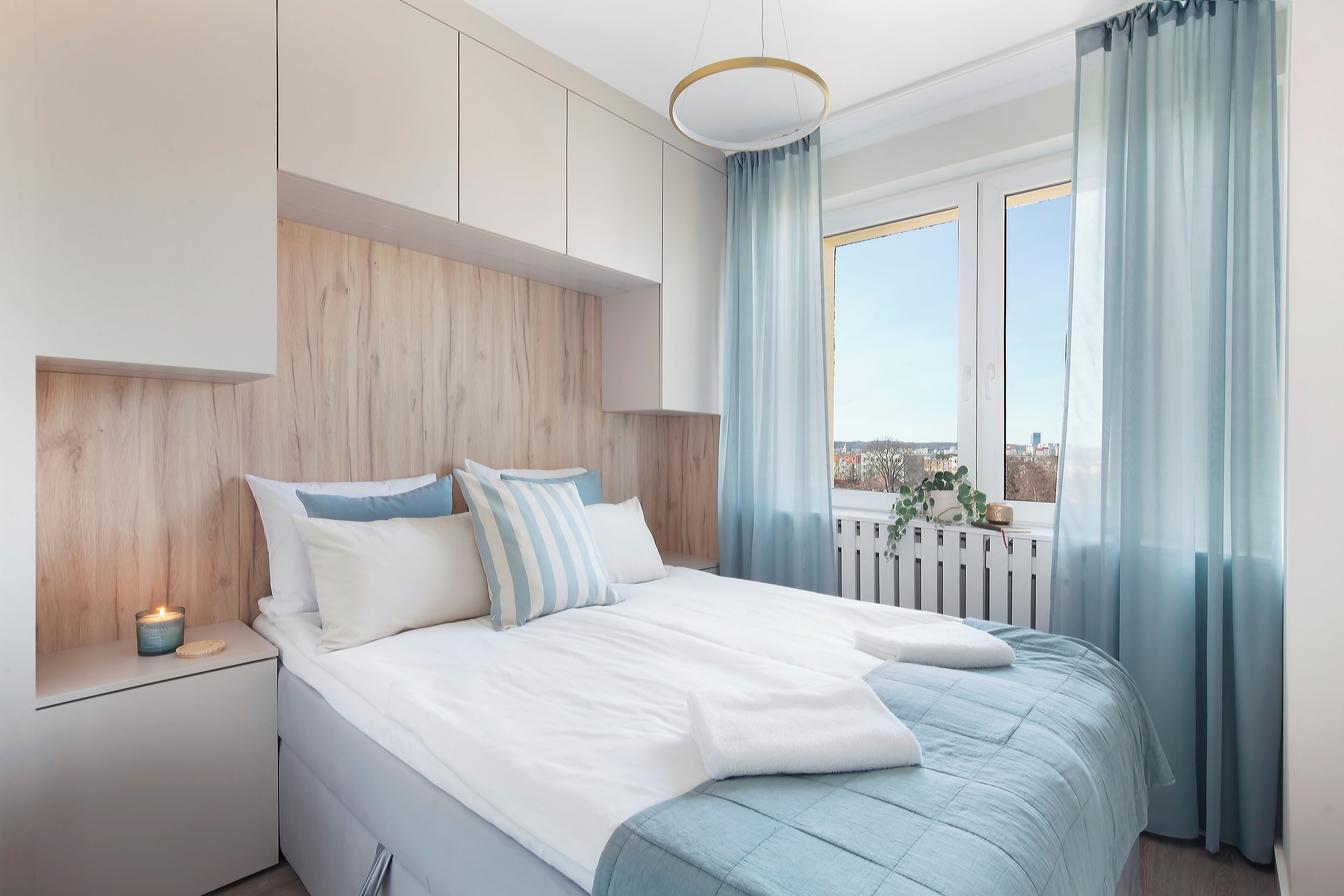 Une chambre moderne avec une tête de lit en bois, un lit blanc avec une literie bleue, une table de chevet avec une bougie, et une vue sur la ville à travers de grandes fenêtres.