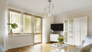 A modern living room with a sofa, a coffee table, and a TV. There are windows with blinds, a plant, and a cabinet with shelves. - Elite Apartments Beachfront (Gdansk)