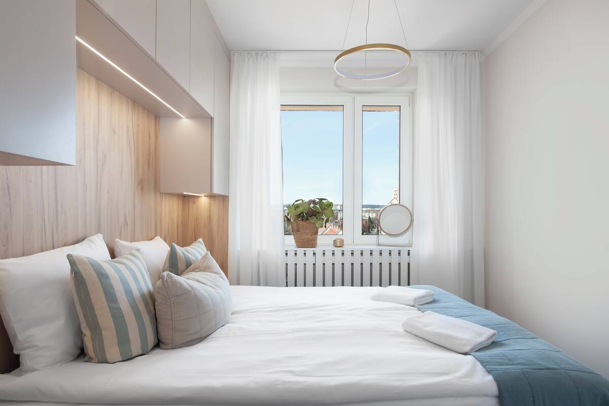 Un dormitorio moderno con una cama grande, ropa de cama blanca, una planta en maceta junto a la ventana y un espejo redondo en la pared.