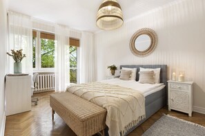 A bedroom with a large bed, a bedside table with candles, a wicker bench, and a view of greenery outside. - Elite Apartments Beachfront (Gdansk)