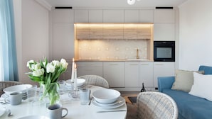 A modern kitchen with white cabinets, a built-in oven, and a dining area with wicker furniture and a vase of white flowers. - Elite Apartments Beachfront (Gdansk)