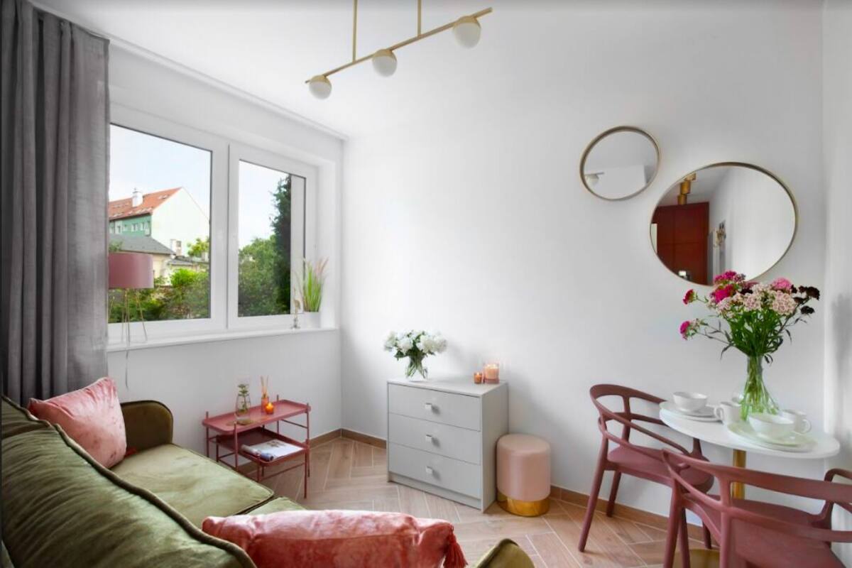 Una habitación moderna con un sofá verde, una mesita con un florero, un espejo redondo y una ventana con vistas a casas.