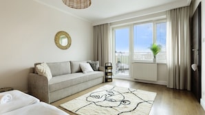 A modern living room with a sofa, a large window with curtains, a hanging light fixture, and a decorative mirror on the wall. - Elite Apartments Beachfront (Gdansk)
