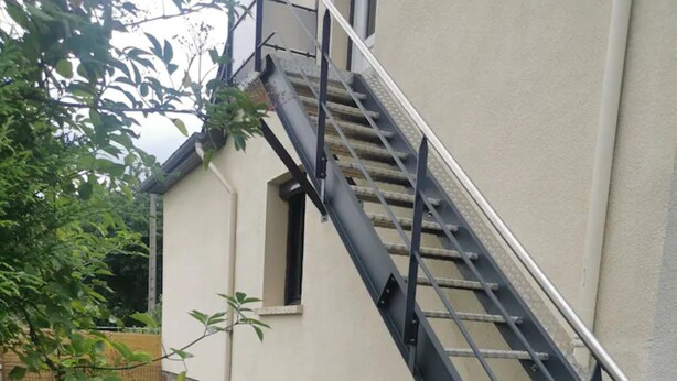 A backyard with a stone wall, a barbecue grill, a table with chairs, and a staircase leading to a second-story window.