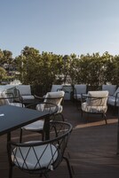Un patio al aire libre con mesas redondas y sillas acolchadas, rodeado de vegetación y con vista a un cuerpo de agua.