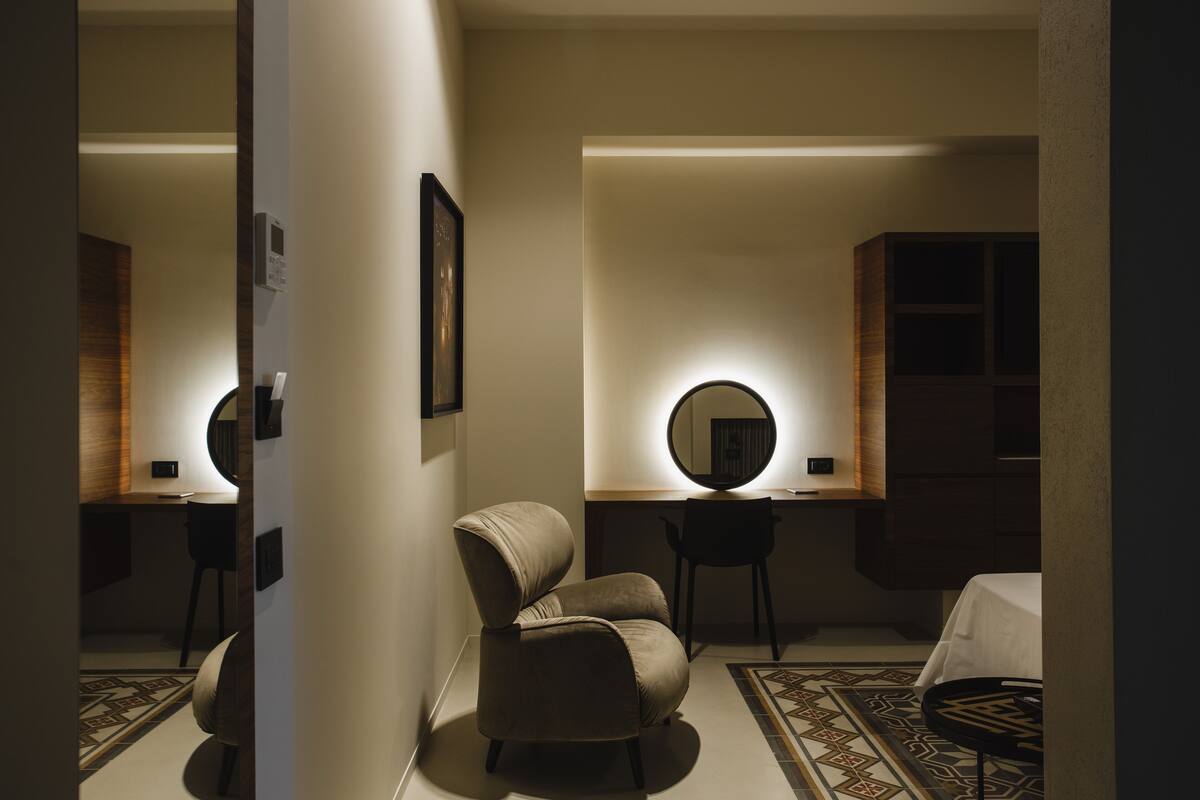 A hotel room with a round mirror, a cushioned armchair, a desk, and a bed with a white bedspread.