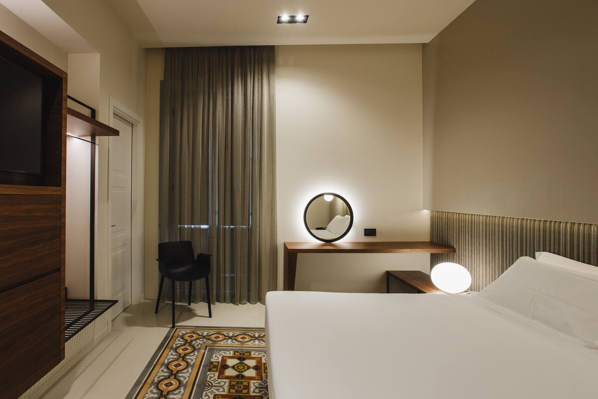 A hotel room with a bed, a desk with a mirror, a chair, and a television.