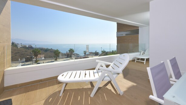 Een balkon met twee witte loungestoelen, een glazen reling en uitzicht op zee en heuvels in de verte.