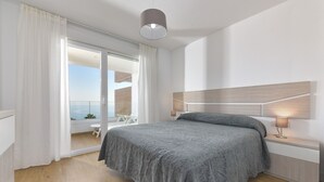 Een slaapkamer met een bed, nachtkastje, lamp, een schuifdeur naar een balkon en uitzicht op de lucht.