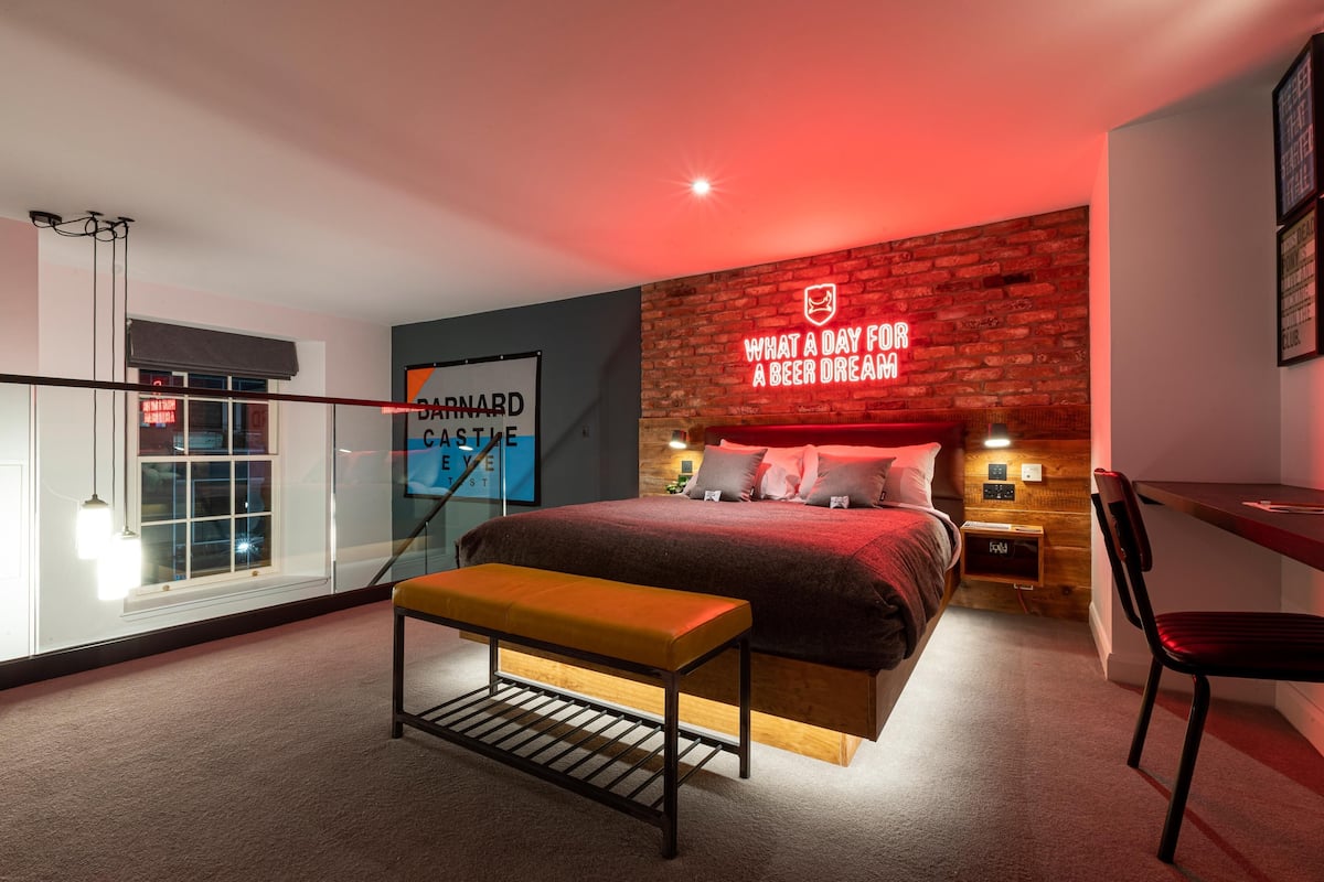 A modern bedroom with a bed, a bench, a desk, and a neon sign on the wall.