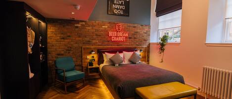 Une chambre d’hôtel avec un mur en briques, un lit recouvert d’une couverture rouge, un banc, une chaise, une table de chevet et une enseigne sur laquelle on peut lire : « BEER DREAM CHARIOT ».