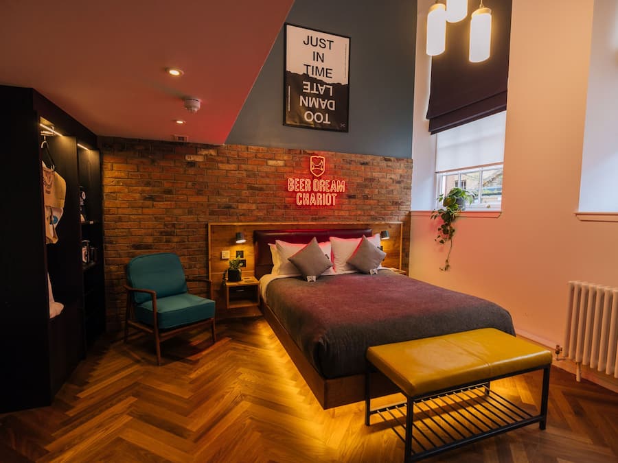Habitación de hotel con pared de ladrillo, cama con manta roja, banco, silla, mesita de noche y un cartel que dice 'BEER DREAM CHARIOT'.