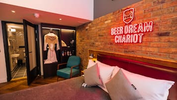 A hotel room with a brick wall, a neon sign reading "Beer Dream Chariot", a bed with pillows, a teal chair, and a robe hanging on a rack.