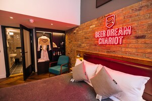A hotel room with a brick wall, a neon sign reading "Beer Dream Chariot", a bed with pillows, a teal chair, and a robe hanging on a rack.