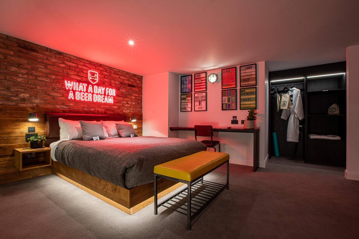 A modern bedroom with a brick wall, a neon sign, a bed, a bench, a desk, and a walk-in closet.