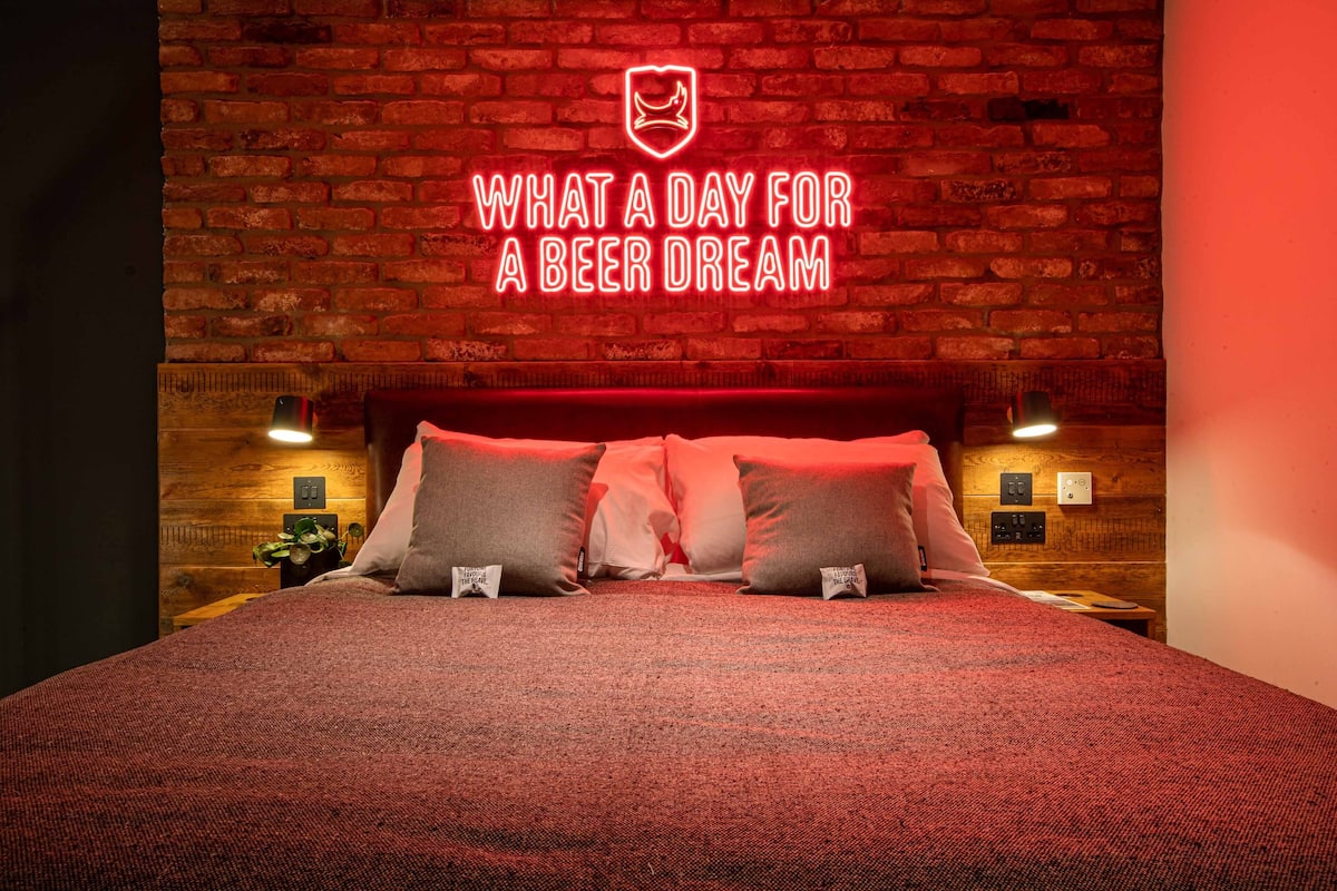 A hotel room with a brick wall, a bed with red bedding, and a neon sign above the bed.