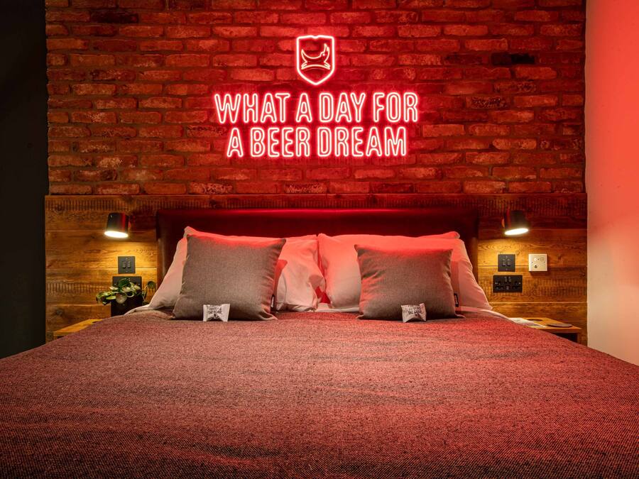 A hotel room with a brick wall, a bed with red bedding, and a neon sign above the bed.