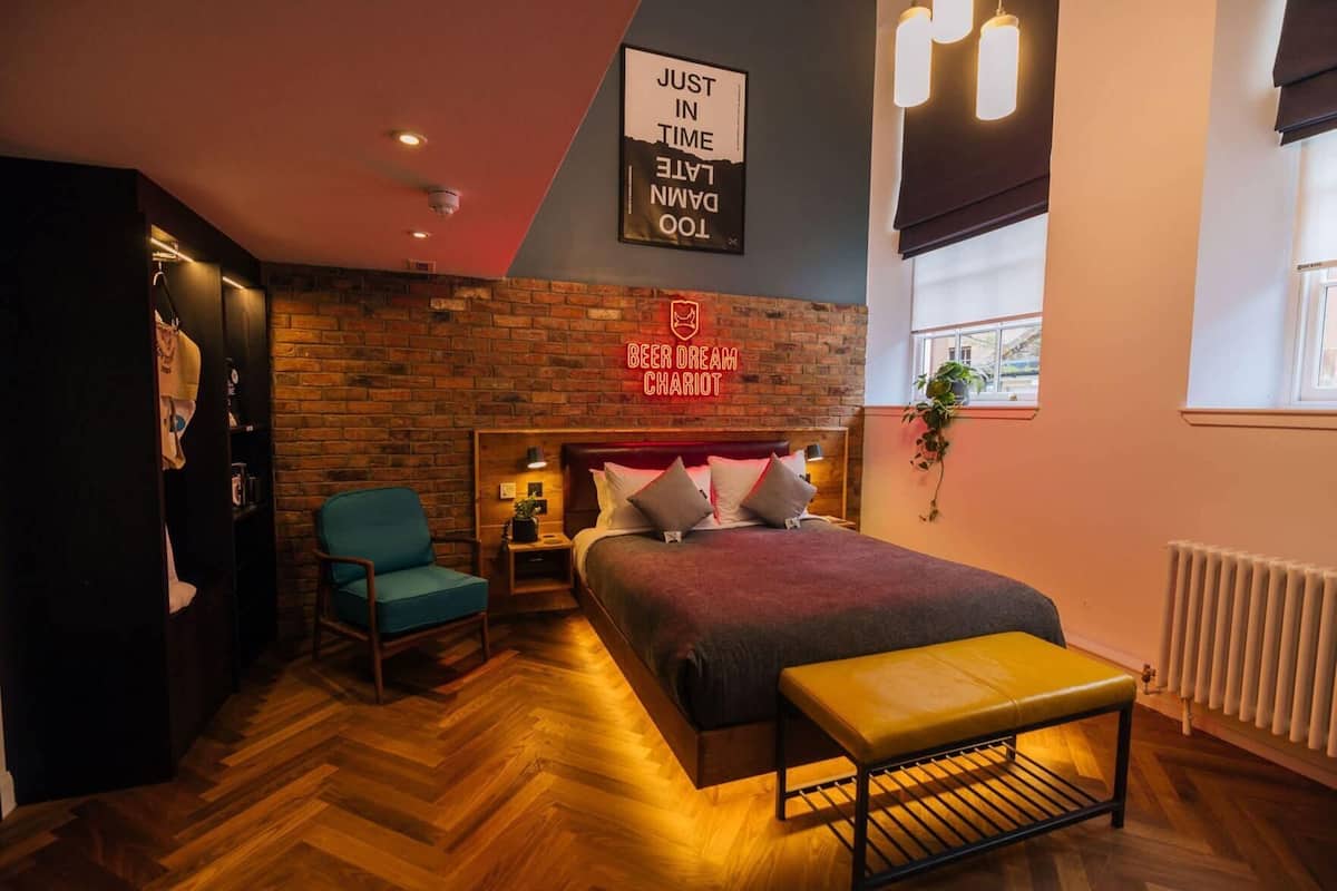 A hotel room with a brick wall, a bed with pillows, a chair, a bedside table, and a window.