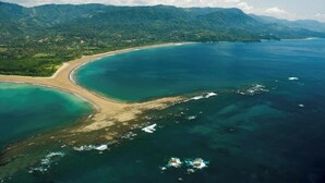 An aerial view of a coastal area with a beach, clear waters, and a mountainous backdrop.