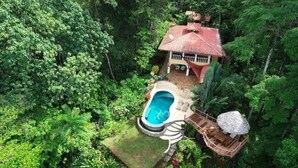 An aerial view of a house surrounded by dense forest, with a swimming pool and a wooden deck.