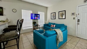 Un salon avec un canapé bleu, un coin repas avec une chaise, un téléviseur fixé au mur et une porte.