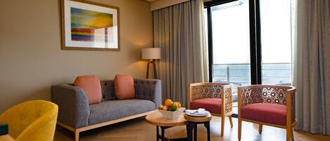 Uma sala de estar moderna com sofá, duas poltronas, uma mesinha com frutas, luminária e uma grande janela com cortinas.