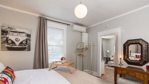 Un dormitorio con cama, mesita de noche, cómoda, espejo, perchero y ventana con cortinas.