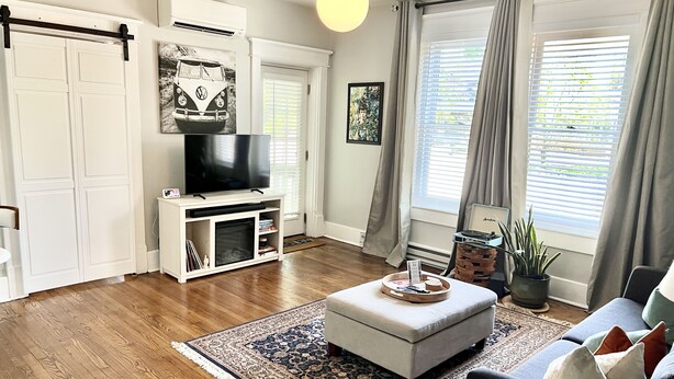 Sala de estar con televisor, reposapiés y sofá. Se ve una ventana con cortinas, una planta y una alfombra.