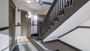 Un pasillo con piso a cuadros, una escalera con barandilla negra y un detector de incendios en la pared.