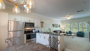 Cozinha moderna com eletrodomésticos de aço inoxidável, mesa de jantar de mármore e sala de estar com sofá e janelas.