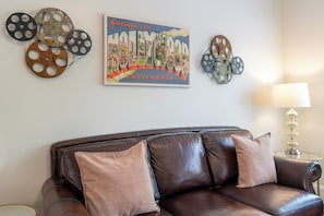 A brown leather sofa with three cushions, a wall-mounted decorative piece with a film reel design, and a framed sign that reads 'Greetings from Hollywood, California.'