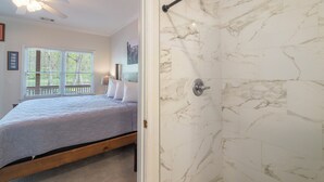 Quarto com cama, chuveiro com paredes de mármore e janela com persianas.