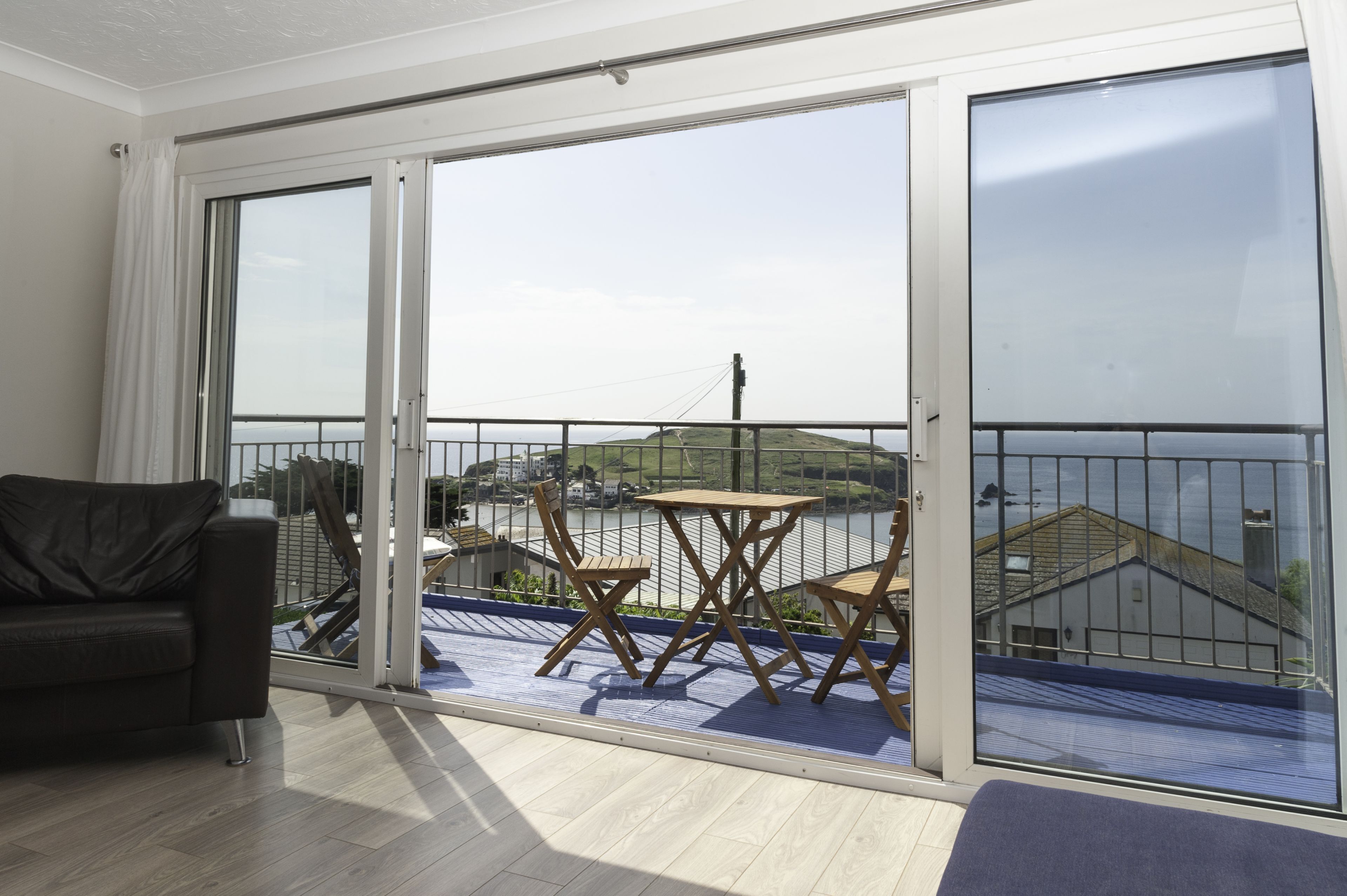 A balcony with a view of a hill and water, a black leather sofa, and a small table with chairs.